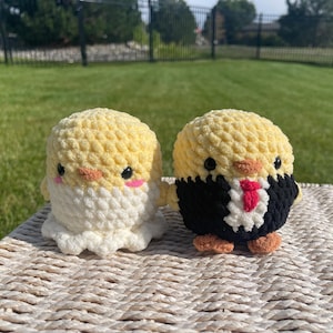 May include: Two handmade crochet chick toys. One chick is dressed in a white dress, and the other is wearing a black suit with a red tie. Both have yellow bodies, black eyes, and pink cheeks. They are sitting on a woven surface.