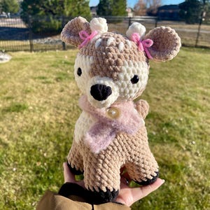 Puede incluir: Un peluche de ciervo hecho a mano con ganchillo en tonos marrón, crema y negro. El ciervo tiene una bufanda rosa y pequeños lazos rosas en las orejas. El peluche se sostiene en una mano.