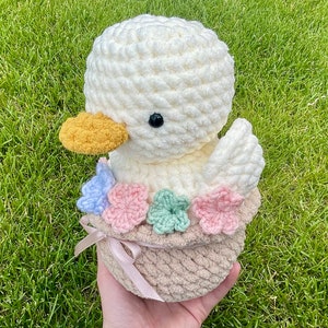 May include: A handmade, crocheted duckling in a basket. The duckling is white with a yellow beak and black eyes. The basket is beige and decorated with pastel-colored flowers and a ribbon.