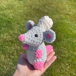 May include: A handmade crochet mouse toy in shades of gray, white, and pink. The plush toy has a gray body, pink ears, feet, and nose, and a white head. The mouse has black eyes and is held in a hand.