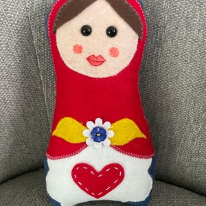 Op de afbeelding: Een handgemaakte vilten pop met een rode jurk, een gele sjerp en een witte bloem met een blauwe knoop in het midden. De pop heeft een wit gezicht met bruin haar, zwarte ogen en rode lippen.