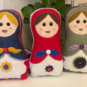 Op de afbeelding: Drie handgemaakte Russische poppen van vilt. De poppen staan in een rij, van links naar rechts: blauw, rood en groen. Elke pop heeft een wit gezicht met geborduurde kenmerken, zwarte ogen en een rode mond. De poppen zijn versierd met gele, blauwe en paarse vilt accenten.