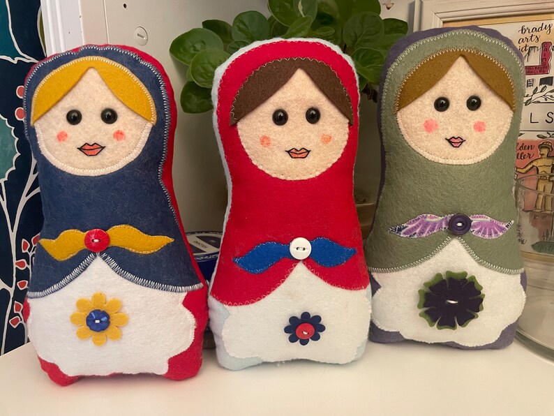 Op de afbeelding: Drie handgemaakte Russische poppen van vilt. De poppen staan in een rij, van links naar rechts: blauw, rood en groen. Elke pop heeft een wit gezicht met geborduurde kenmerken, zwarte ogen en een rode mond. De poppen zijn versierd met gele, blauwe en paarse vilt accenten.