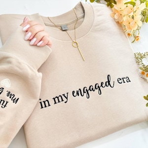 May include: A beige sweatshirt embroidered with the words "in my engaged era" in black script. The left sleeve features "Future Mrs. Brown" embroidery. A gold necklace and flowers are also present.