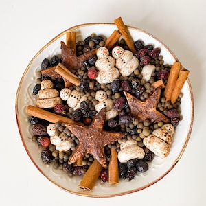 Może przedstawiać: Biała ceramiczna miska wypełniona różnymi składnikami potpourri, w tym patyczkami cynamonu, anyżem gwiazdkowym, suszonymi jagodami i małymi figurkami bałwanów.