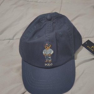 May include: A blue baseball cap featuring an embroidered graphic of a bear wearing a denim shirt and a cap. The word POLO is embroidered in white thread below the bear. A black tag with the text RALPH is partially visible on the right side.