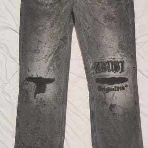 May include: Dark gray denim jeans with distressed details, including a ripped knee and a patch with the text "RSWT Original 1999". The jeans have a faded, textured appearance and a brown tag attached.