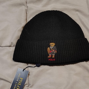 May include: Black knit beanie featuring an embroidered bear wearing a suit. The beanie has a folded brim and a small tag attached. The bear is wearing a suit and holding a bag. The word "POLO" is embroidered on the hat.