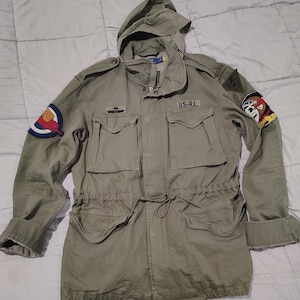 May include: Olive green military-style jacket with a hood, multiple pockets, and a drawstring waist. Features patches on the sleeves, including a red and blue emblem and a cartoon character. The text "U.S.-R.L." is visible.