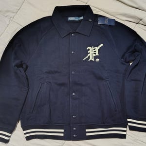 Puede incluir: Chaqueta universitaria azul marino con botones, con una gran "P" estilizada en color crema claro en el pecho. La chaqueta tiene rayas blancas en los puños y el dobladillo, y un pequeño logotipo en la manga izquierda.