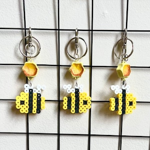 May include: Three yellow and black beaded bumble bee keychains with a honeycomb charm. Each keychain is attached to a silver key ring.