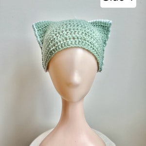 Puede incluir: Un gorro de punto de color verde claro con ribete blanco, con forma de orejas de gato. El gorro está en una cabeza de maniquí blanca. Lado 1.