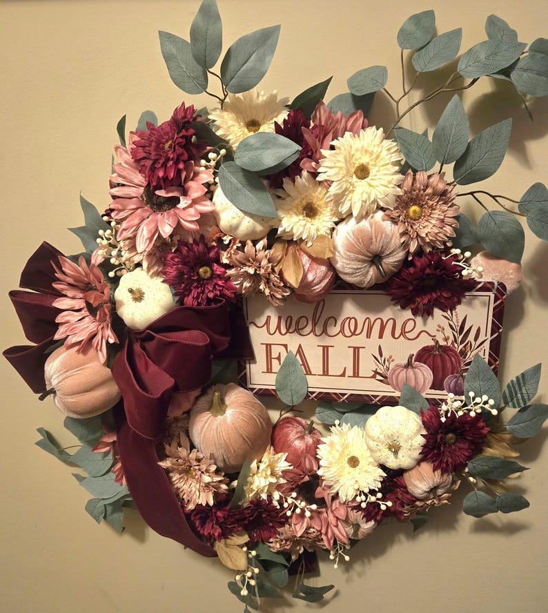 May include: A fall wreath with burgundy, pink, and white flowers, greenery, and velvet pumpkins. A sign with the text 'welcome fall' is in the center of the wreath.