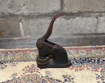 May include: A vintage metal seal stamp with a black finish and a rusty brown handle. The stamp has a circular design and is sitting on a patterned rug.