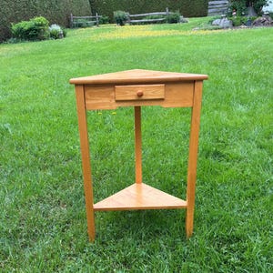 May include: A light brown wooden corner table featuring a triangular top, three straight legs, a single drawer with a small round knob centered on the front apron, and a lower triangular shelf positioned between the legs for additional storage space.