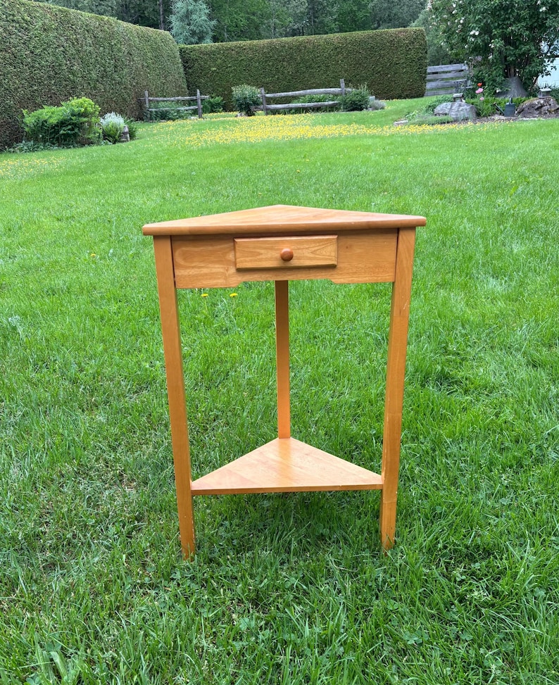 May include: A light brown wooden corner table featuring a triangular top, three straight legs, a single drawer with a small round knob centred on the front apron, and a lower triangular shelf positioned between the legs for additional storage space.