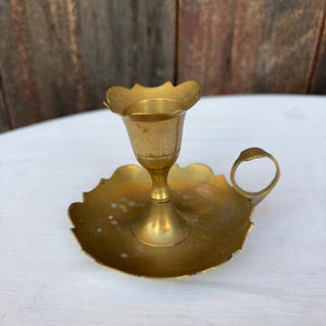 Puede incluir: Un candelero de latón vintage con una copa en forma de flor y una base en forma de hoja. El candelero tiene un asa pequeña y está hecho de un material de latón brillante y envejecido. El soporte está diseñado para una sola vela.