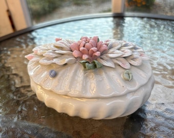 Puede incluir: Una caja de porcelana blanca con un diseño floral. La caja tiene forma de concha y presenta una flor rosa y blanca con hojas verdes.