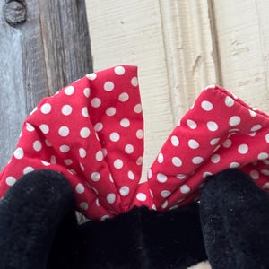 May include: A close-up of a plush toy featuring a red bow with white polka dots. The bow is positioned above black plush ears and a cream-colored face. The background is a weathered wooden surface.
