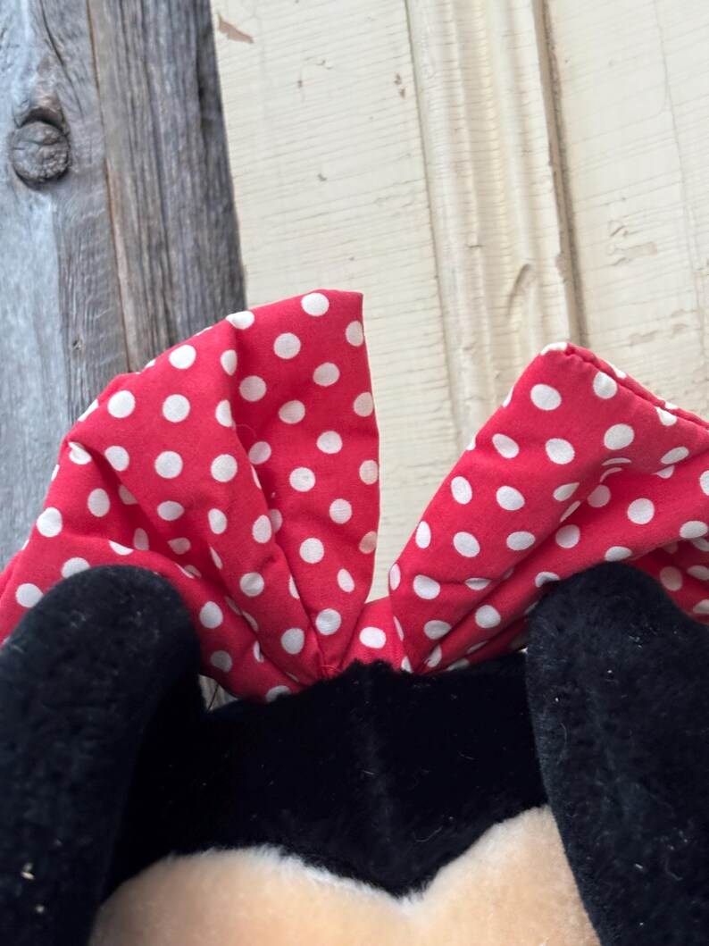 May include: A close-up of a plush toy featuring a red bow with white polka dots. The bow is positioned above black plush ears and a cream-colored face. The background is a weathered wooden surface.
