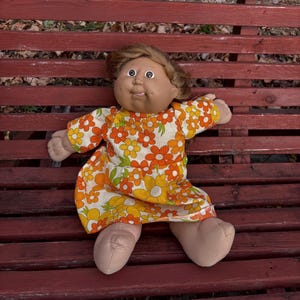 Puede incluir: Una muñeca vintage con tez bronceada, cabello castaño y ojos azules. La muñeca lleva un vestido con estampado floral con flores naranjas, amarillas y blancas. La muñeca está sentada en un banco de madera rojo.