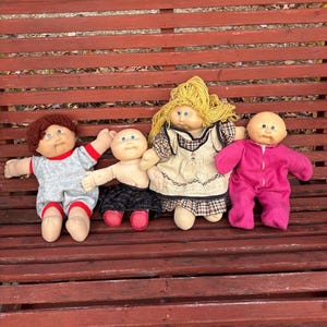 Puede incluir: Cuatro muñecas vintage Cabbage Patch Kids. Una muñeca viste un atuendo gris y rojo, otra una falda negra y zapatos rojos, una tercera un vestido y la última un mono rosa. Las muñecas están dispuestas sobre un banco de madera rojo.