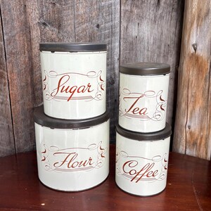 May include: A set of four cylindrical metal canisters with dark brown lids and cream-coloured bodies. Each canister features decorative brown script text reading Sugar, Flour, Tea, and Coffee, accented with ornate swirling lines on the front surface.