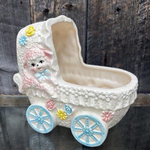 Könnte beinhalten: Vintage Babywagen-Blumentopf aus cremefarbener Keramik mit blauen und rosa Akzenten. Der Wagen hat eine Lammfigur, Blumendetails und blaue Räder. Ein charmantes Dekorationsstück.