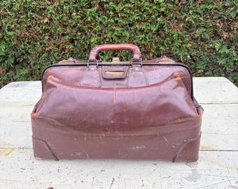 May include: A dark brown leather doctor bag with a structured rectangular frame, a top handle, and a metal clasp closure. The exterior shows signs of wear with visible scuffs and creases on the surface, featuring reinforced corners and stitching.
