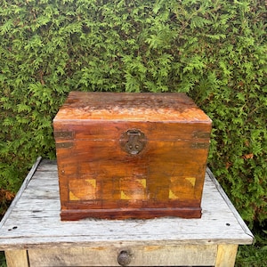 May include: A rectangular wooden chest with a polished brown finish, featuring a central metal latch mechanism and decorative gold-toned square accents along the bottom front panel, showcasing a visible wood grain texture throughout the exterior.
