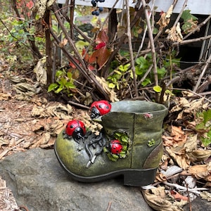 May include: A green, weathered boot-shaped planter, adorned with three red ladybug decorations. The boot has a distressed finish and sits on a grey stone. The planter is surrounded by foliage, suggesting a garden theme.