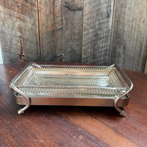 Peut inclure: Un plateau rectangulaire en verre transparent avec un motif de bordure perlée texturée, inséré dans un cadre en métal argenté poli. Le cadre comporte quatre pieds incurvés qui surélèvent le plateau en verre, formant un support de présentation.