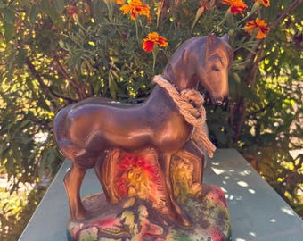 May include: A brown horse figurine with a rope around its neck, standing on a colorful base. The horse is set against a backdrop of orange flowers and green foliage, creating a rustic aesthetic.