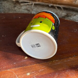 May include: A white ceramic mug featuring a cartoon illustration of a green-skinned character with orange hair on a black background with white dots. The bottom of the mug displays two small square icons indicating dishwasher and microwave safety.