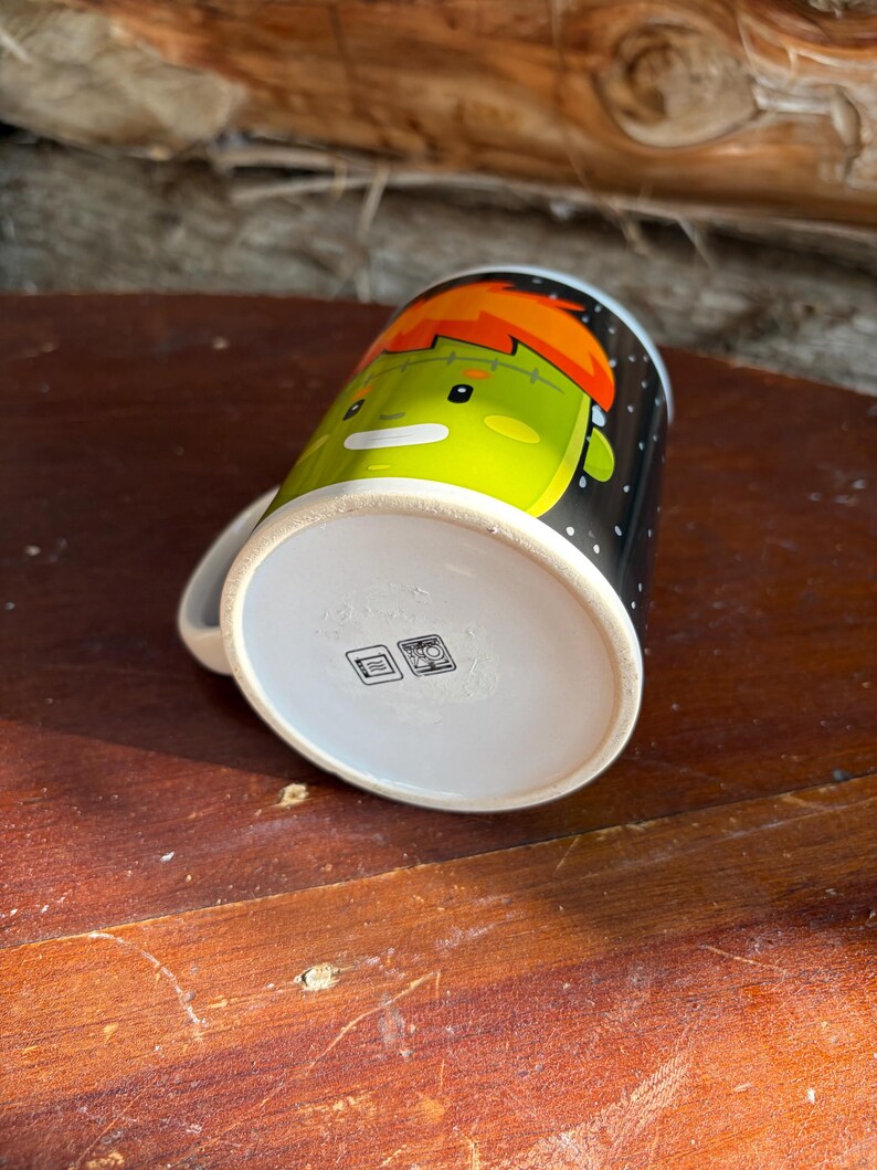 May include: A white ceramic mug featuring a cartoon illustration of a green-skinned character with orange hair on a black background with white dots. The bottom of the mug displays two small square icons indicating dishwasher and microwave safety.