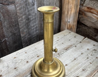 May include: A vintage brass candlestick holder with a round base and a tall, cylindrical stem. The candlestick has a small knob and a flared top. The brass has a warm, aged patina, suggesting it is an antique or vintage piece.