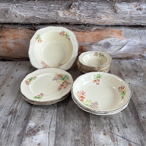 May include: A collection of vintage cream-coloured ceramic dishware. The set includes plates, bowls, and serving dishes, each adorned with a floral pattern in shades of orange, green, and purple. The edges of the dishes have a gold trim.