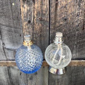 Puede incluir: Dos lámparas de aceite decorativas. Una es azul con un patrón de diamantes, la otra es transparente con un efecto craquelado. Ambas tienen detalles plateados y dorados y una mecha. Las lámparas son redondas y están sobre una superficie de madera.