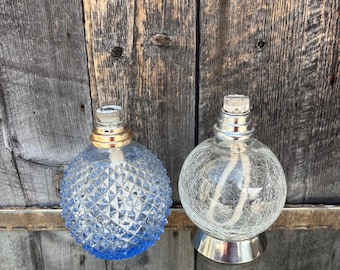 Könnte beinhalten: Zwei dekorative Öllampen. Eine ist blau mit einem Rautenmuster, die andere ist klar mit einem Krakelierungseffekt. Beide haben silberne und goldene Akzente und einen Docht. Die Lampen sind rund und stehen auf einer Holzoberfläche.