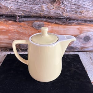 Puede incluir: Una tetera de cerámica de color amarillo pálido con tapa a juego y boquilla blanca. La tetera tiene un asa curva y se encuentra sobre una superficie negra. El fondo es una pared de madera.