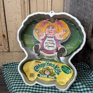Puede incluir: Un molde para pastel vintage de Cabbage Patch Kids. El molde es de color plateado y tiene la forma de una muñeca Cabbage Patch Kid. El embalaje incluye una imagen de la muñeca con el texto "Happy Birthday Lauren". El molde está diseñado para hornear un pastel con la forma de la muñeca.