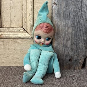 Puede incluir: Una muñeca vintage con un atuendo de terciopelo verde azulado y un gorro puntiagudo a juego. La muñeca tiene una cara pintada con grandes ojos azules, labios rojos y cabello rosado. La muñeca está sentada con las piernas extendidas.