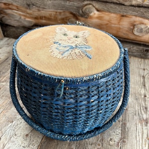 Pode incluir: Uma cesta de vime azul com uma tampa redonda que apresenta um desenho de ponto de agulha de um gato branco com um laço azul. A cesta tem duas alças tecidas.