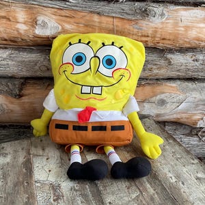 May include: A yellow plush toy of Spongebob Squarepants, a popular cartoon character. The toy is wearing a white shirt with a red tie and brown trousers. The toy has a big smile and blue eyes.