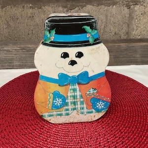 Puede incluir: Una lata vintage con forma de muñeco de nieve con sombrero de copa negro, pajarita azul y chaqueta naranja. El muñeco de nieve tiene una cara sonriente con ojos negros y boca de carbón. La lata está decorada con acebo y copos de nieve.