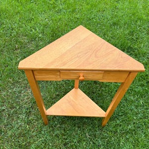 May include: A triangular wooden side table with a light brown finish. The table features a flat top surface, a single drawer with a small round knob centred on the front panel, and a lower triangular shelf supported by three straight legs.