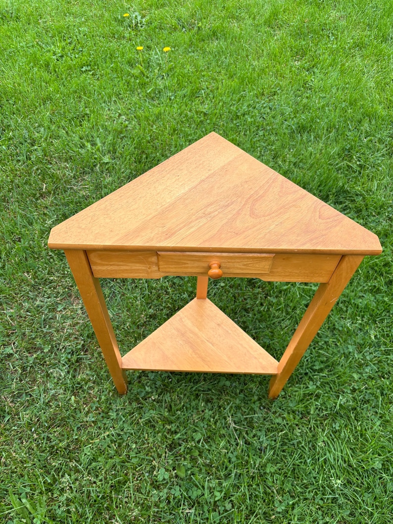 May include: A triangular wooden side table with a light brown finish. The table features a flat top surface, a single drawer with a small round knob centred on the front panel, and a lower triangular shelf supported by three straight legs.