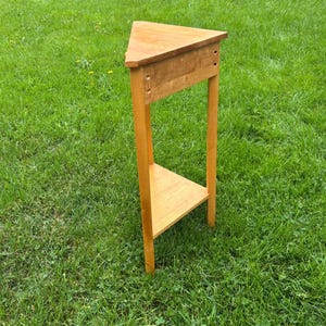 May include: A light brown wooden corner table featuring a triangular top and a matching triangular lower shelf. The table is supported by three vertical legs and includes a horizontal apron connecting the legs beneath the top surface.