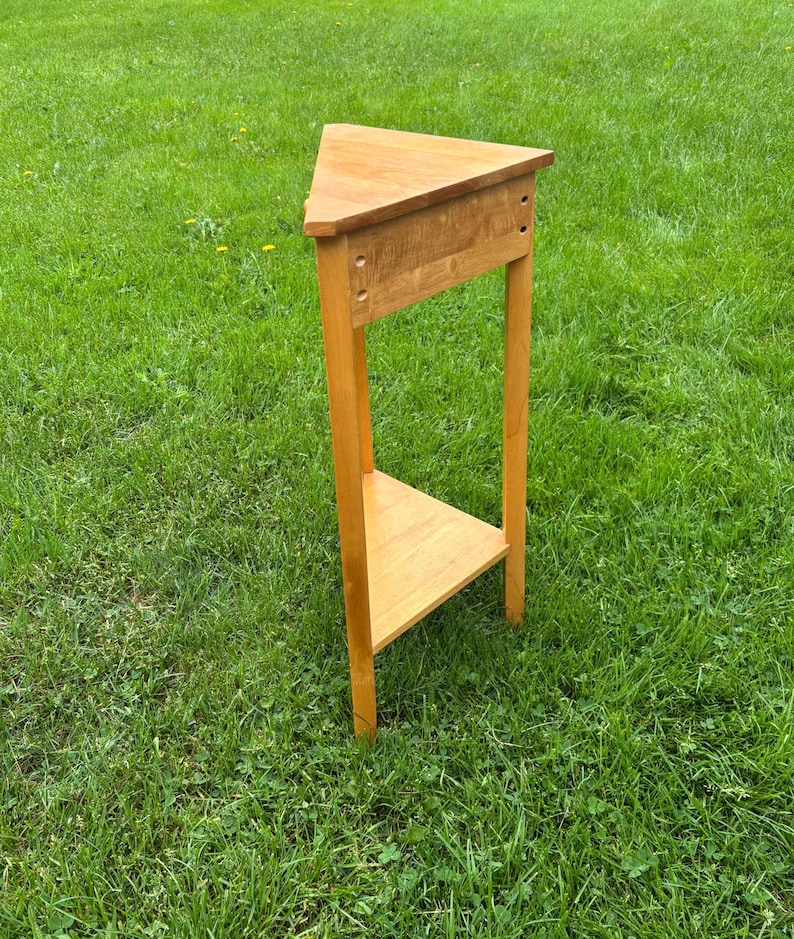 May include: A light brown wooden corner table featuring a triangular top and a matching triangular lower shelf. The table is supported by three vertical legs and includes a horizontal apron connecting the legs beneath the top surface.