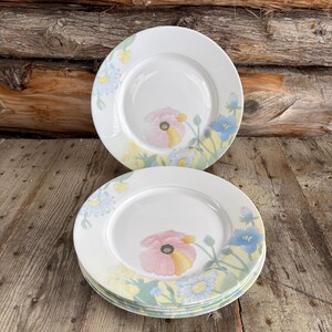 May include: A set of white ceramic plates with a floral pattern. Each plate has a pink poppy and blue and yellow flowers. The plates are on a wooden surface with a wooden backdrop.